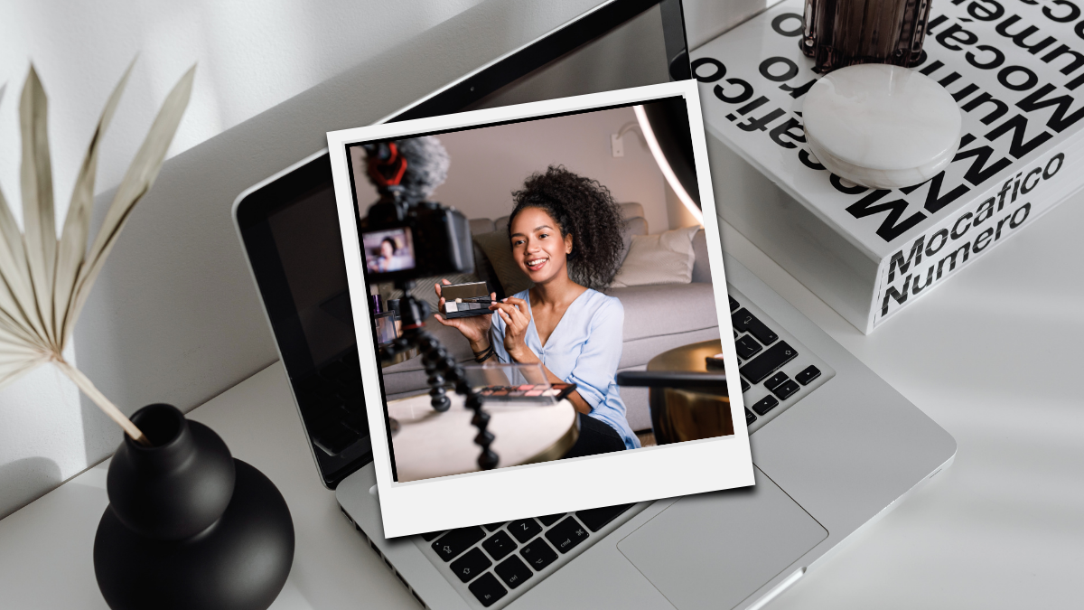 A content creator with curly hair smiles while holding a makeup palette and recording herself on a camera mounted on a tripod, with a Polaroid-style photo overlay on a laptop and desk setup.