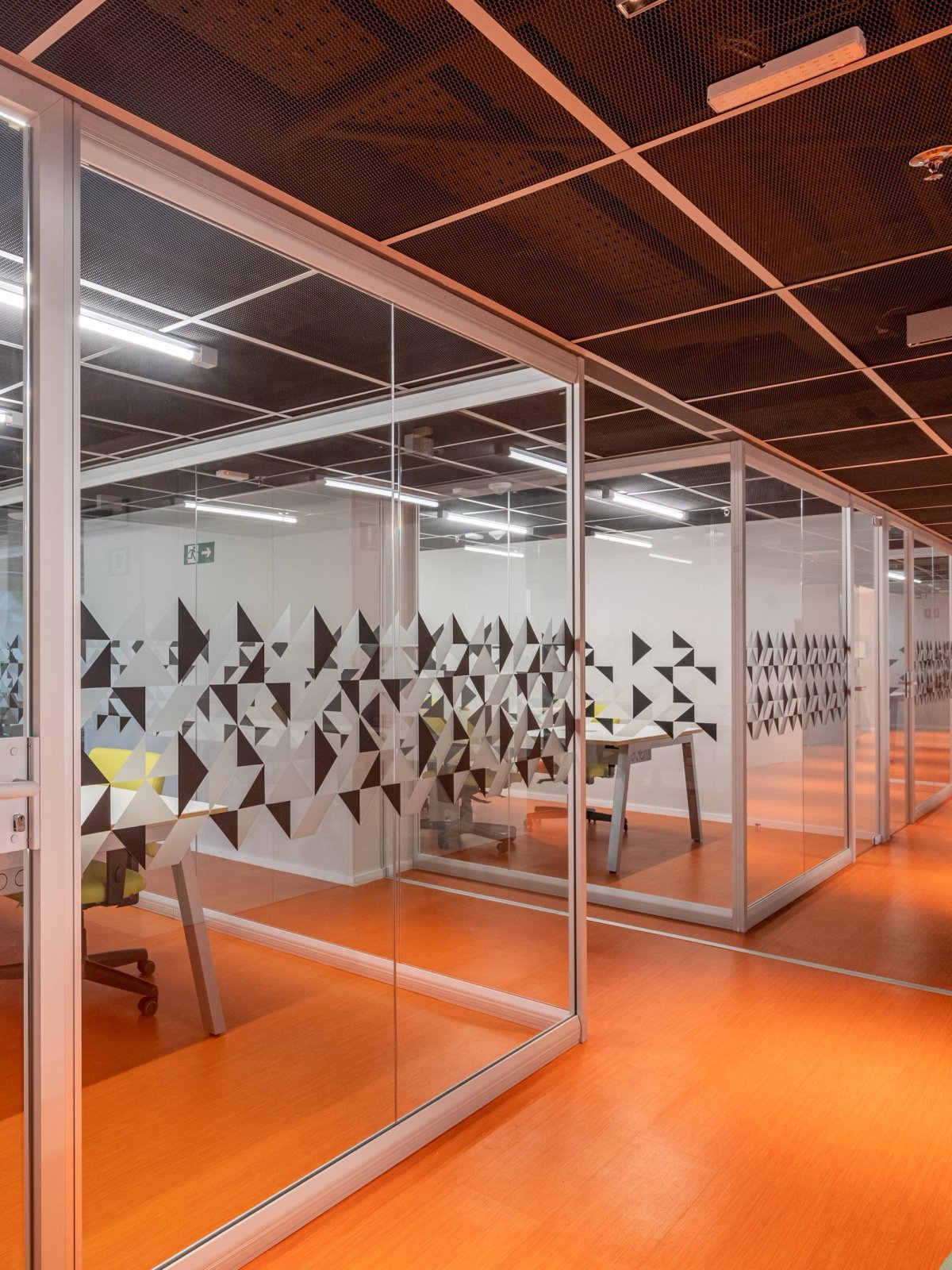Divisórias de vidro da linha Ackua com adesivos geométricos e piso laranja.