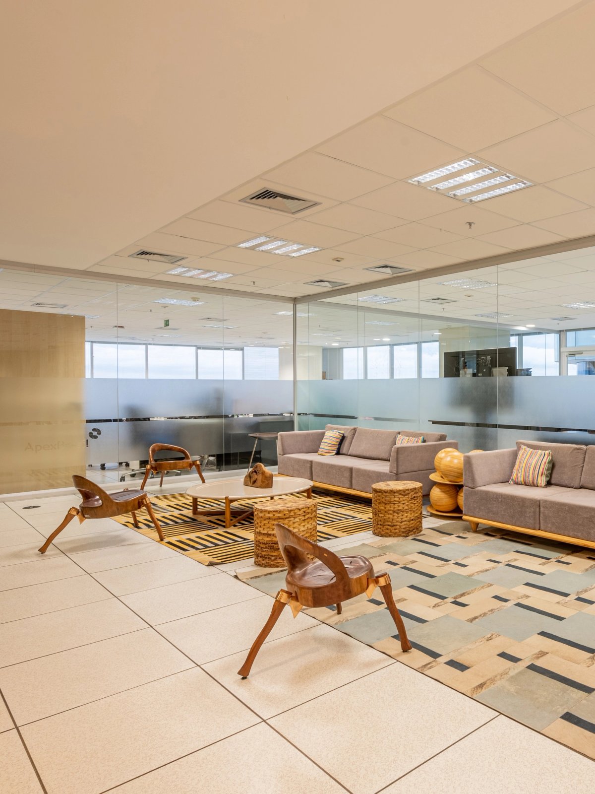 Lounge corporativo com sofá e poltronas de madeira, divisórias de vidro ao fundo.