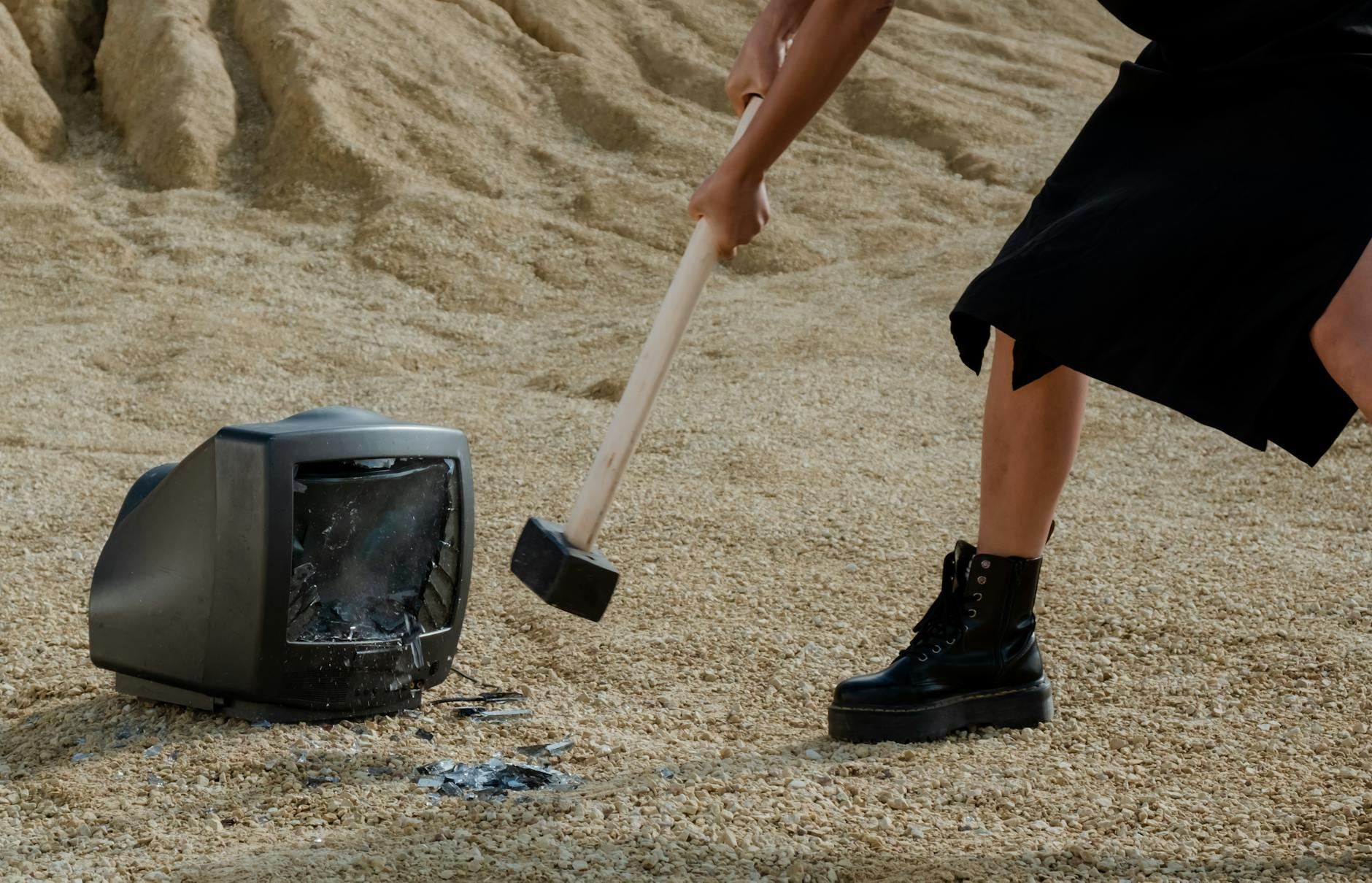 A person in black boots and a dress fiercely smashes an old television with a mallet on sandy ground.