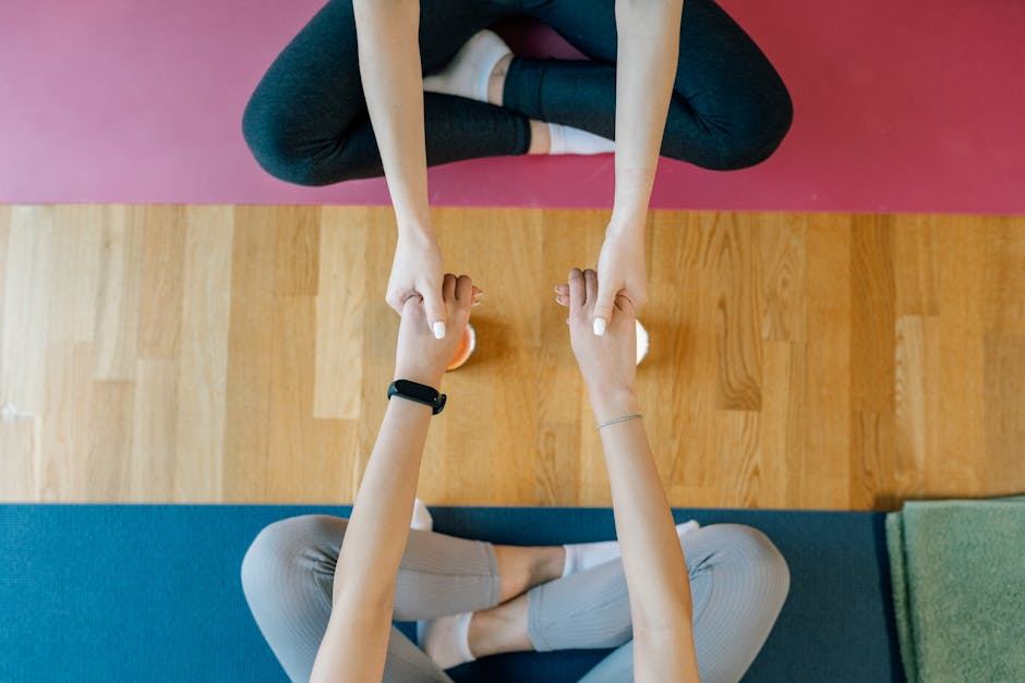 Two individuals are seated on yoga mats, facing each other, with their legs crossed. They are reaching out to hold hands, encouraging connection. The background features a wooden floor, and there are additional mats in different colors nearby. This scene suggests a focus on mindfulness and self-care activities.
