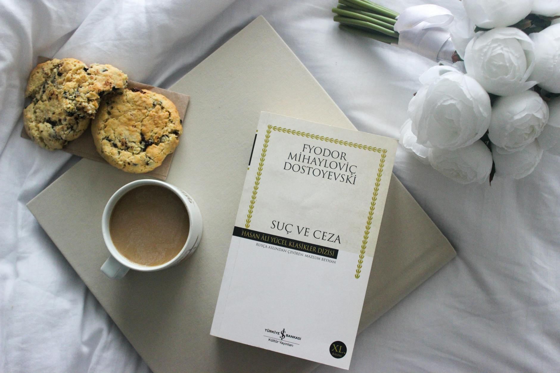 A cozy scene featuring an astrology book, a steaming cup of coffee, and cookies, surrounded by delicate white roses on a bed.