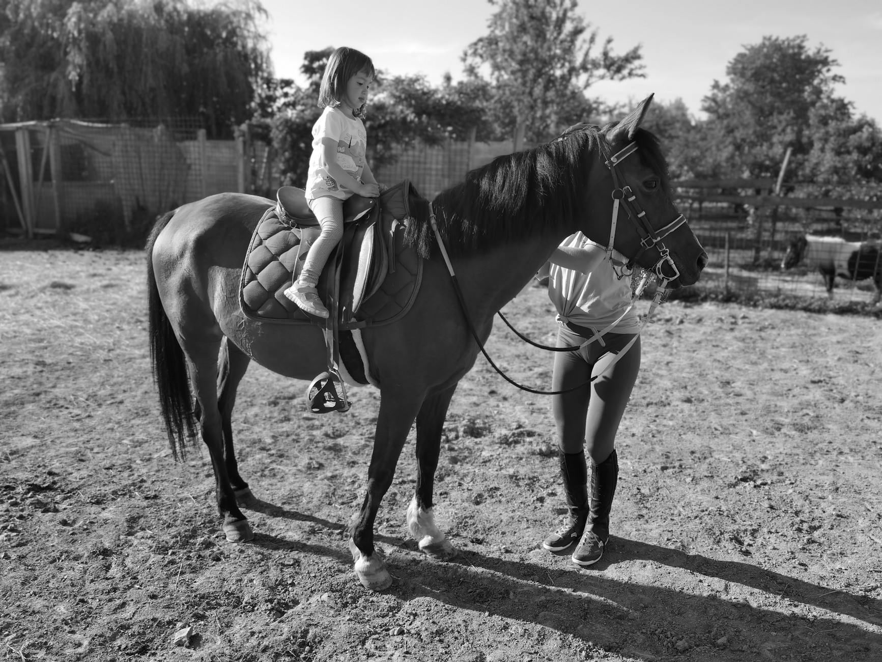 A young girl joyfully rides a pony while a caring woman assists her on a rustic farm.