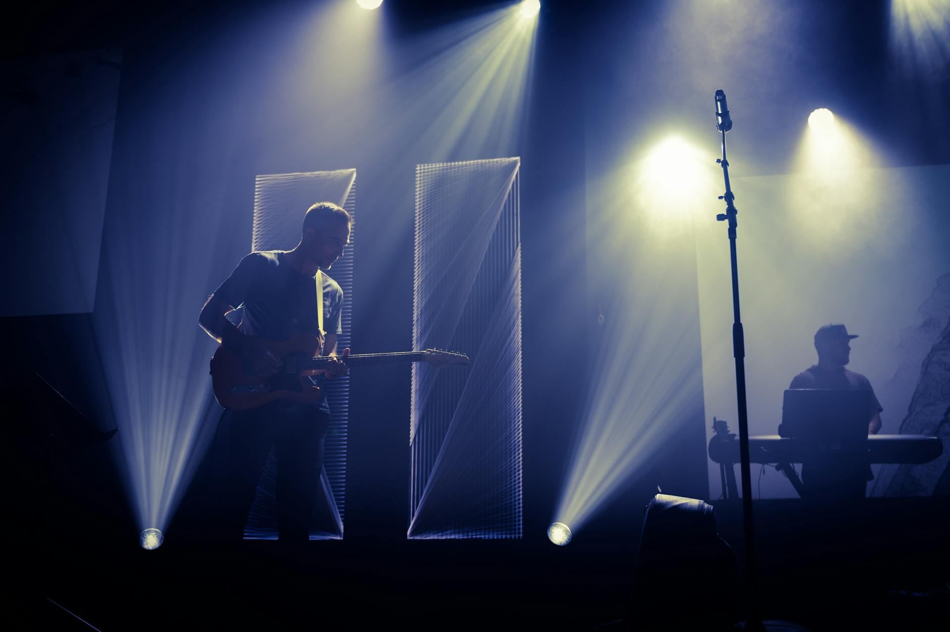 Anonymous male musician performing melody with guitar on stage near microphone and DJ in dark club with spotlights