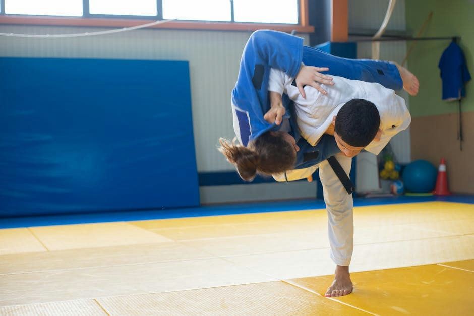 A young person in a white judo gi is performing a stretching pose on a colorful tatami mat, which is yellow and red. The background features large windows allowing natural light to illuminate the space, highlighting the gym's atmosphere. The individual appears focused and engaged in their practice.