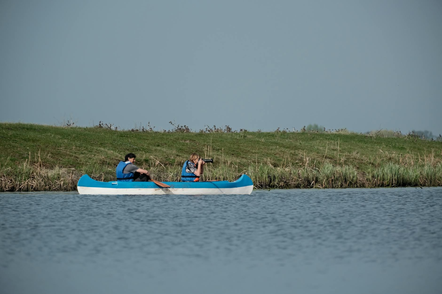 Two adults canoeing on a tranquil lake near grassy shore under a clear sky.