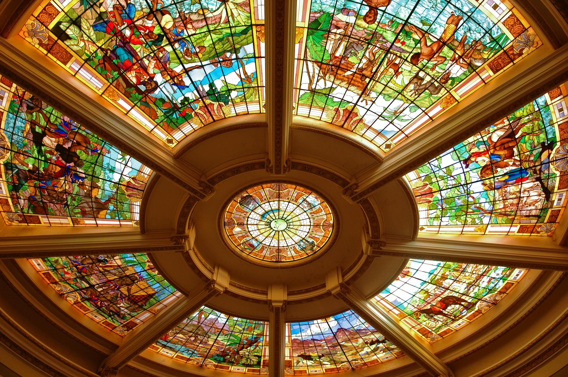 Colorful stained glass dome with intricate artwork, showcasing historical and cultural depictions.