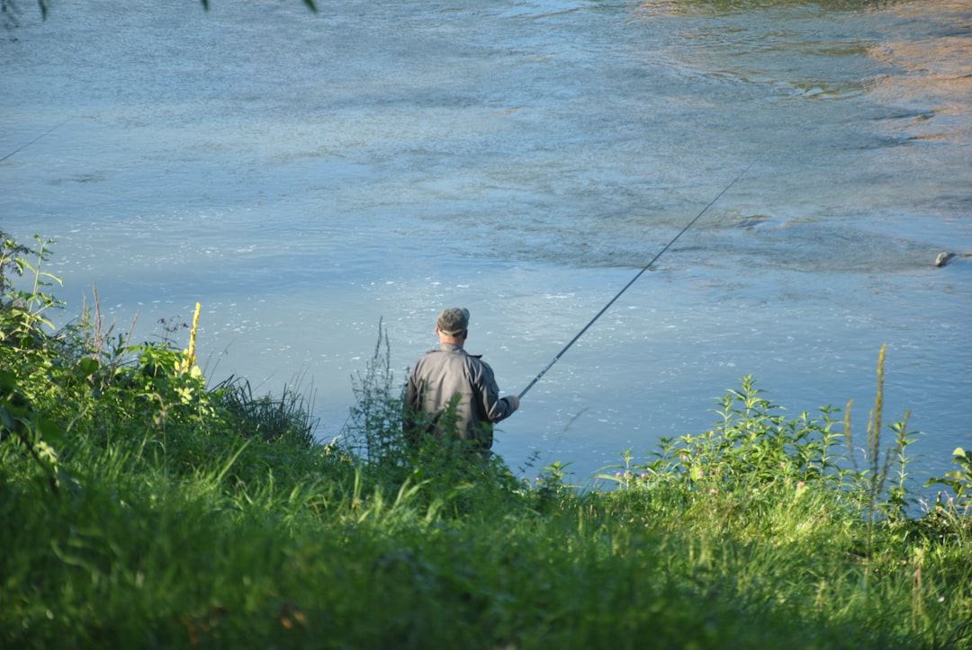 Man fishing on a riverbank with green foliage.