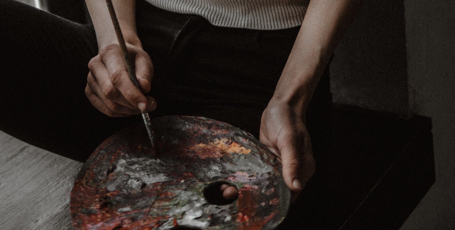 A close-up of hands holding a messy paint palette with various colors, alongside a paintbrush ready for use. The background is softly blurred, creating a cozy artistic atmosphere.