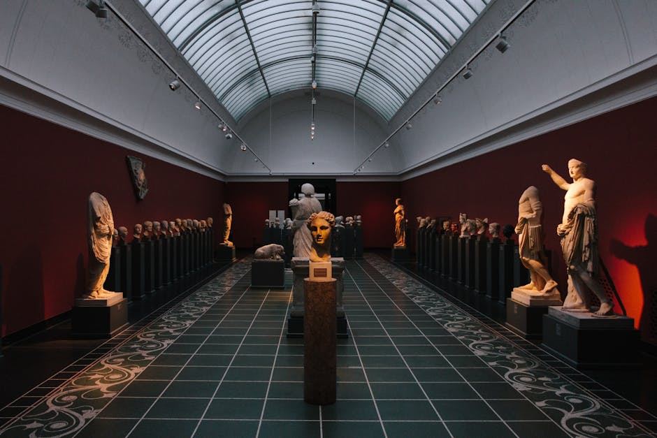 The image shows a spacious museum hall with a high glass ceiling, illuminating a collection of classical sculptures. Statues of various historical figures line the walls, while a central pedestal displays a prominent head sculpture. The dark red walls contrast with the light-colored sculptures, creating a striking visual effect.