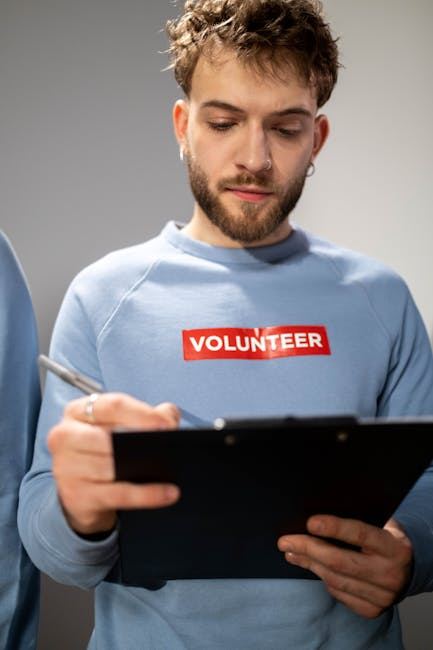 A young man in a blue sweatshirt with 'VOLUNTEER' writes on a clipboard, suggesting involvement in volunteering.