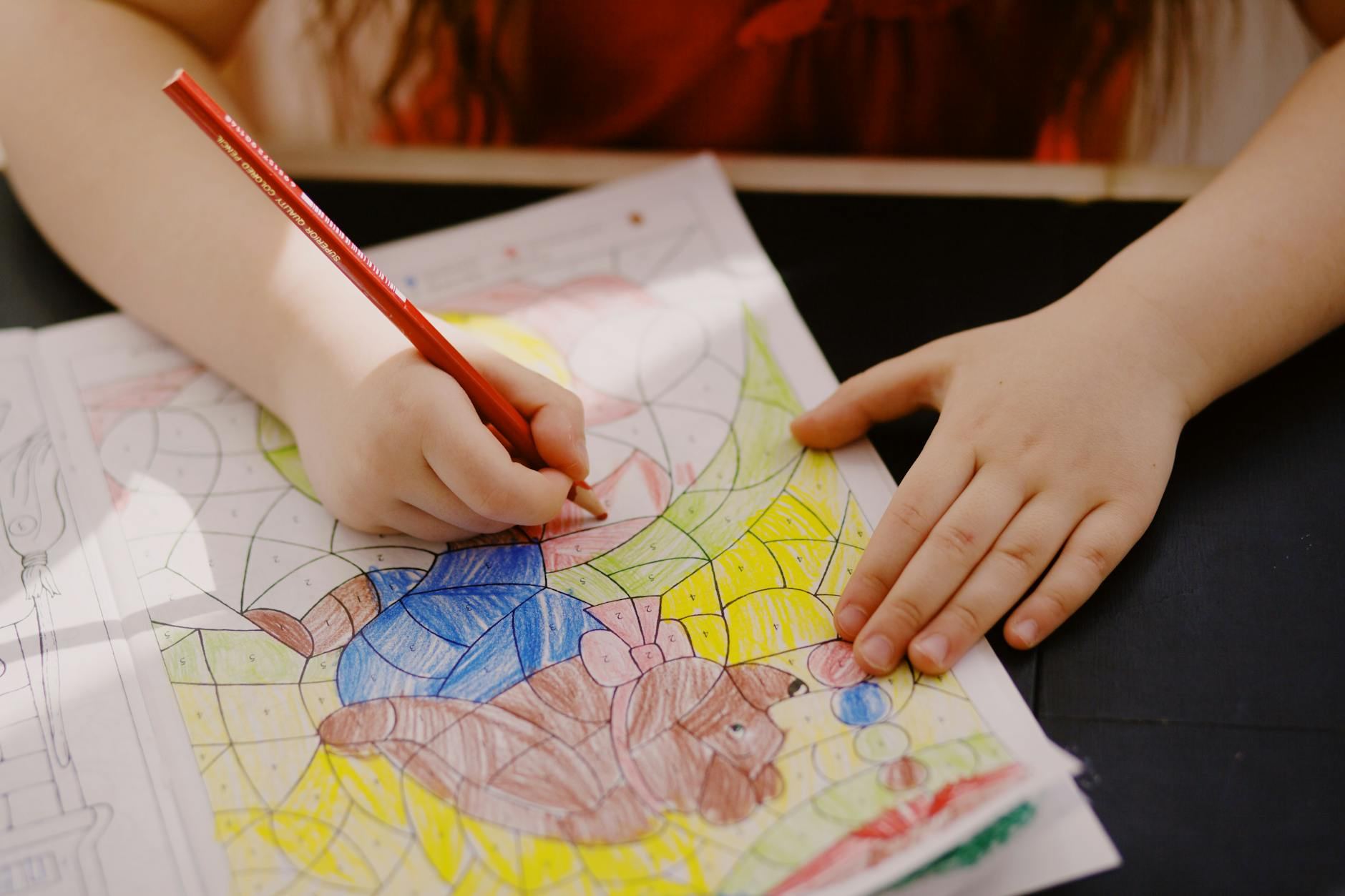 Child drawing with colored pencil on a paper indoors, showcasing creativity and learning.