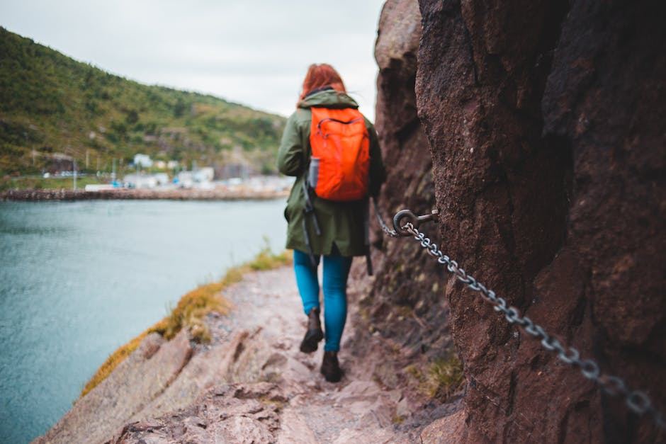 Person in a green coat and orange backpack walking on a rocky trail near a body of water.