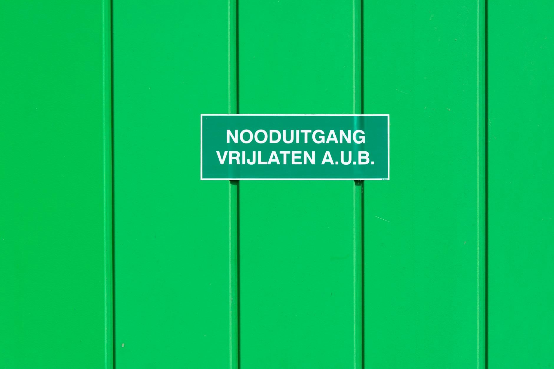 A green metal wall with a bright emergency exit sign in Dutch, emphasizing safety in a paintball arena.