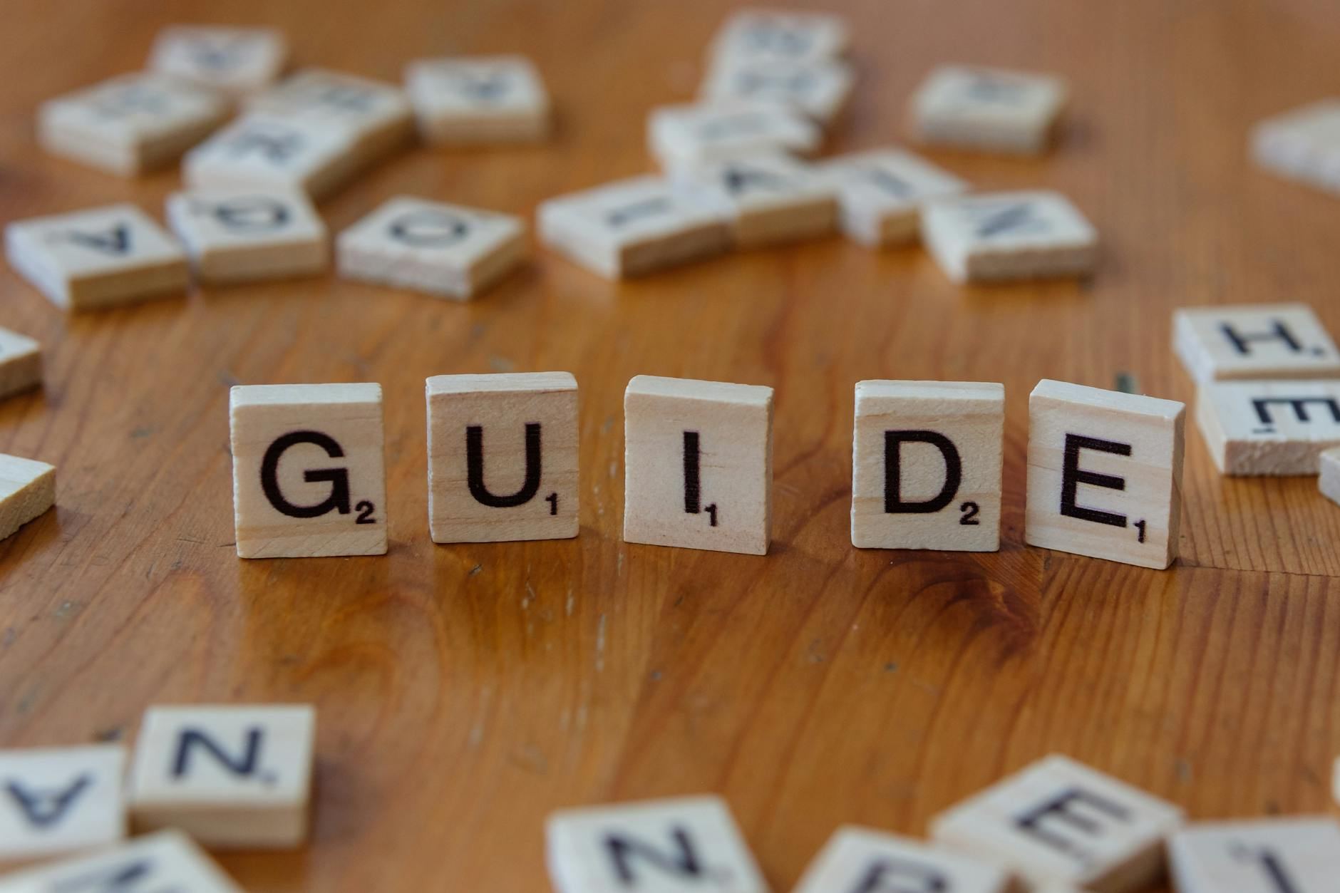 Scrabble tiles spell 'Guide' amidst a colorful array of scattered letters on a rustic wooden table.