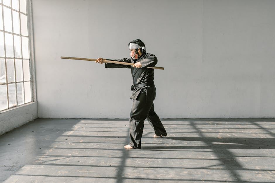 A martial artist dressed in a black gi practices Ninjutsu in a spacious, sunlit room. They hold a wooden staff, demonstrating a dynamic pose. The sunlight casts long shadows on the floor, highlighting the practitioner's focused expression and the smooth movements of their form.