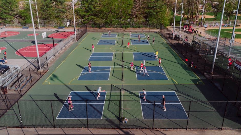 Aerial view of multiple tennis courts with players, surrounded by a basketball court and greenery.