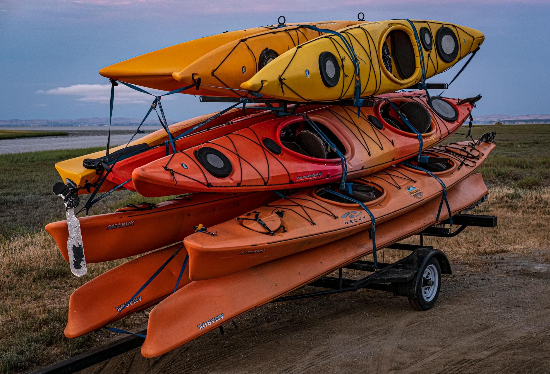 Brightly colored kayaks are neatly stacked on a trailer, set against a picturesque backdrop of lush greenery and blue skies.