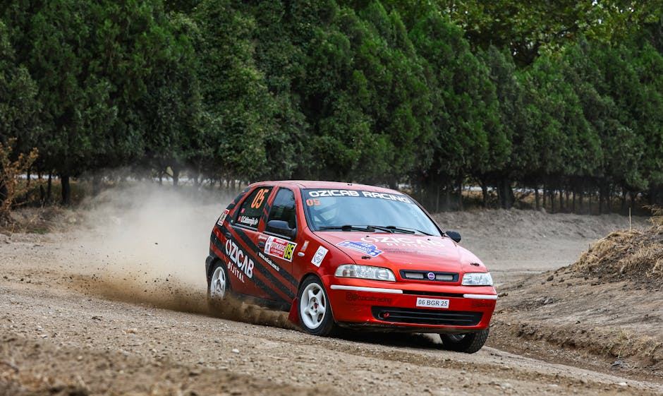 A red rally car kicking up dirt while racing on a gravel track surrounded by trees.