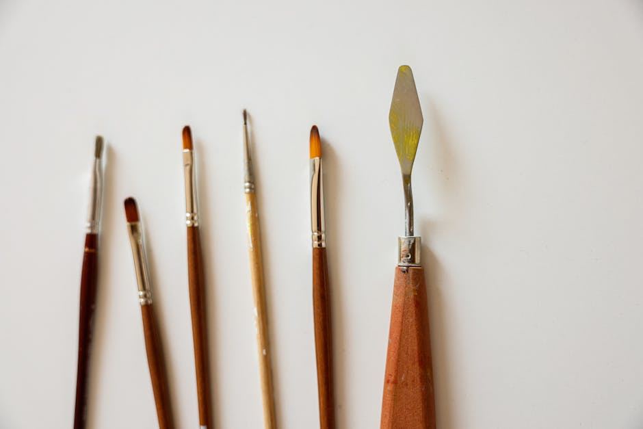 The image features a collection of oil painting tools arranged neatly on a plain white surface. There are five paintbrushes of varying sizes, each with wooden handles and metal ferrules. Next to the brushes, there is a palette knife with a silver blade and a wooden handle, ideal for mixing and applying paint.