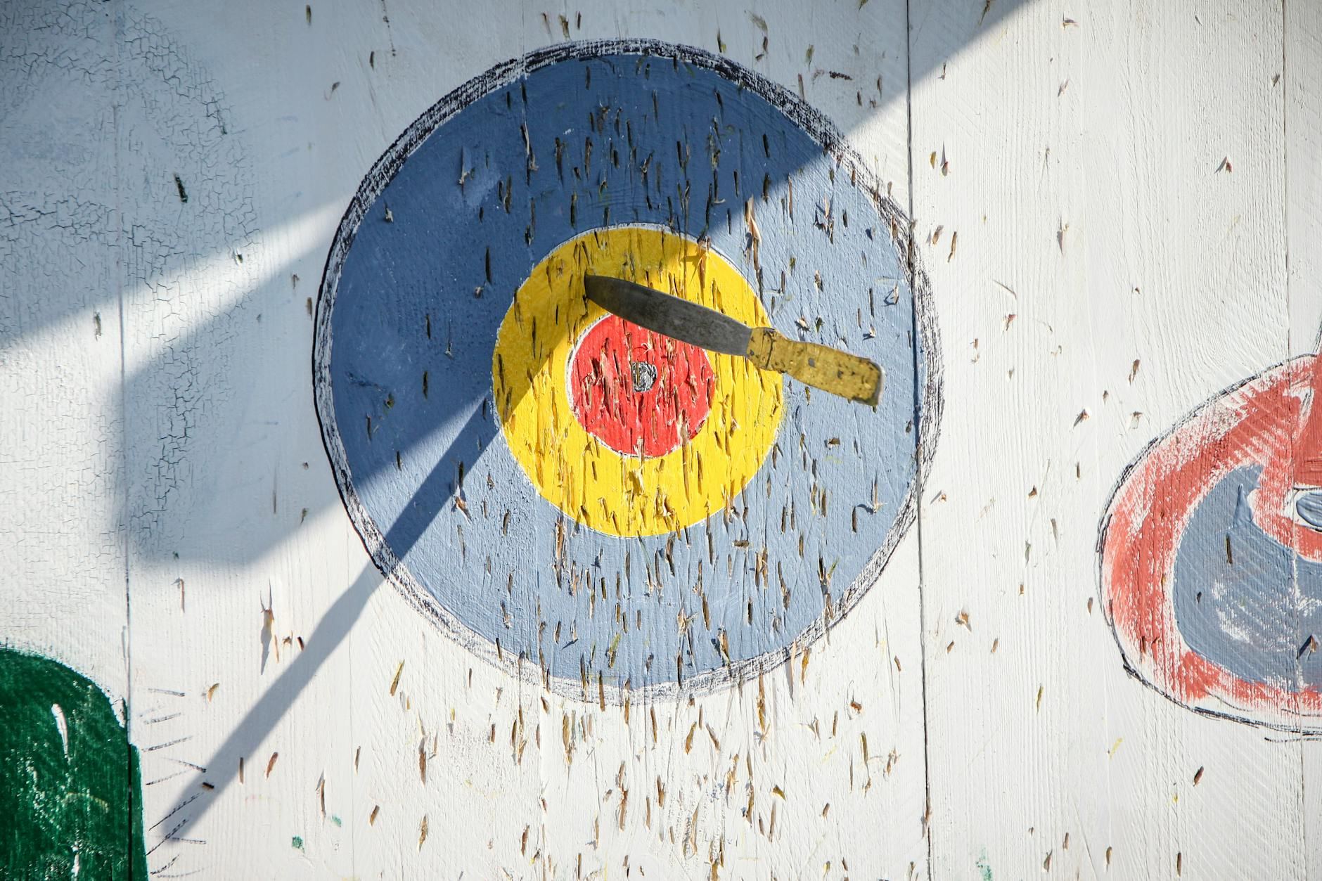 Close-up of a knife stuck in a painted bullseye on a wooden target board during daylight.