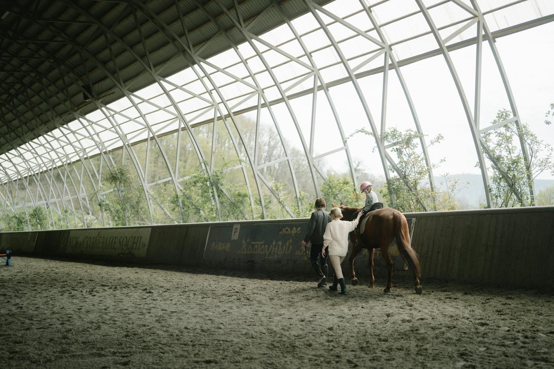 A young child practices horseback riding in a spacious indoor arena, receiving supportive guidance from attentive instructors.