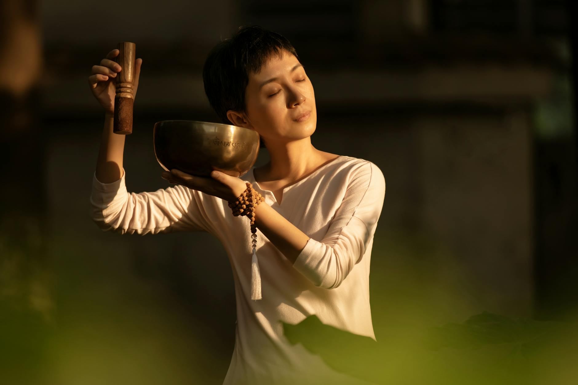 A serene woman sits cross-legged, gently holding a singing bowl, surrounded by soft light, embodying tranquility and mindfulness.
