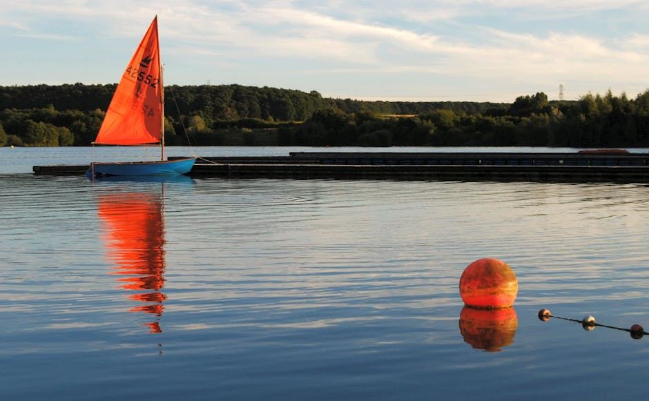 A colorful dinghy sailboat on calm water, reflecting the sky and surrounded by a buoy.