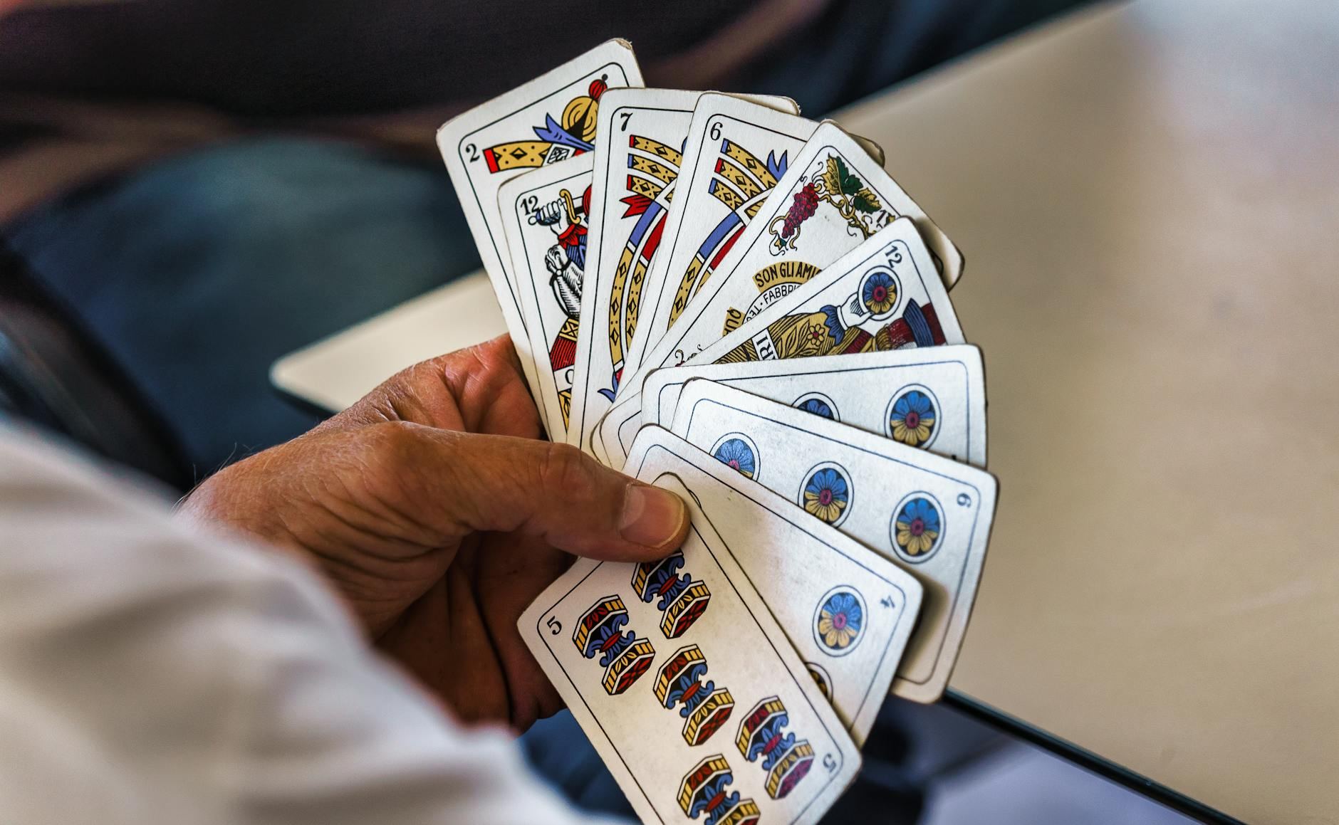 A hand holds a fanned-out set of colorful playing cards displaying various designs and numbers on a tabletop, indicating a card game in progress. The background features blurred seating.