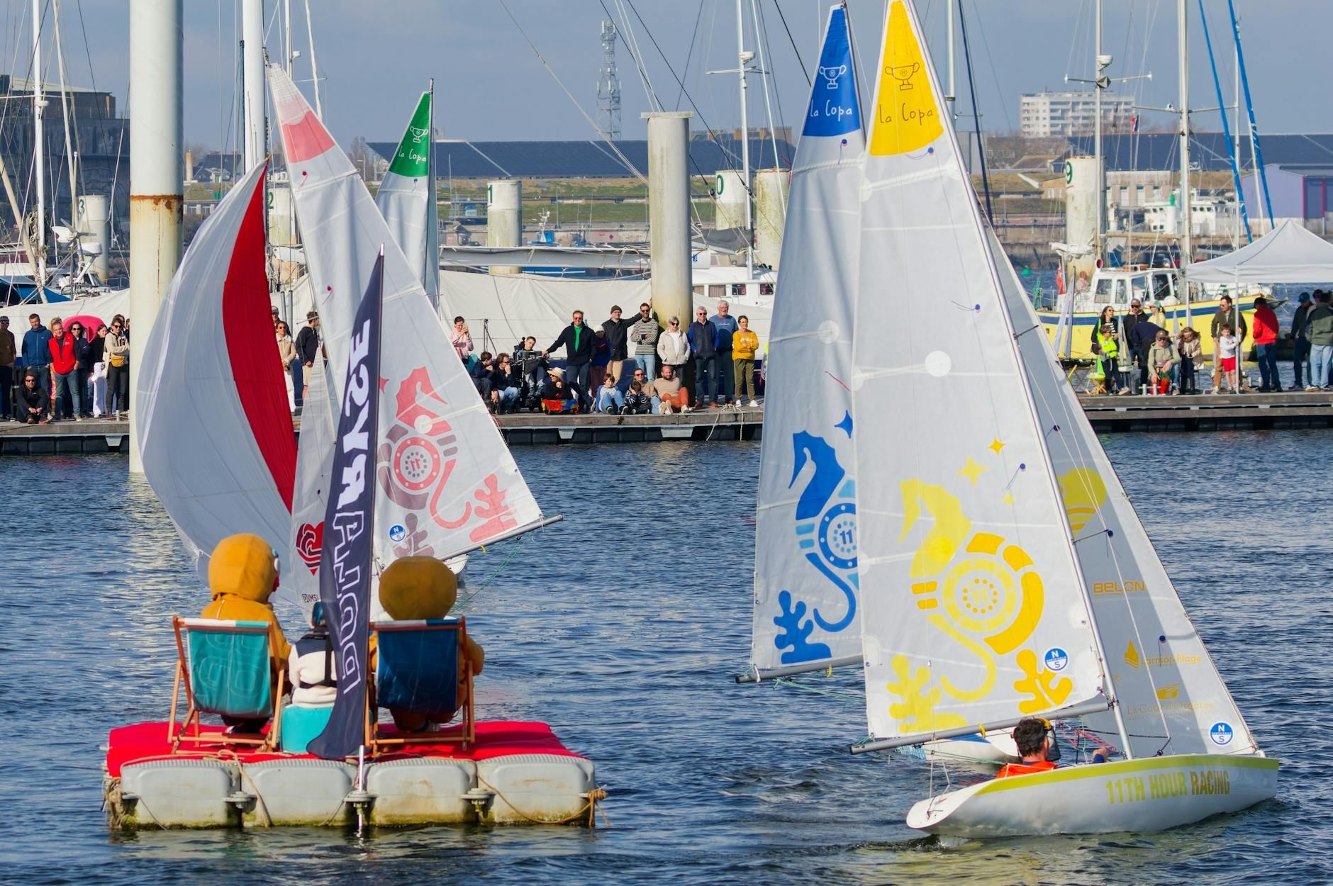 A lively sailing competition at Larmor-Plage features colorful vessels racing amidst an enthusiastic crowd cheering on the participants.