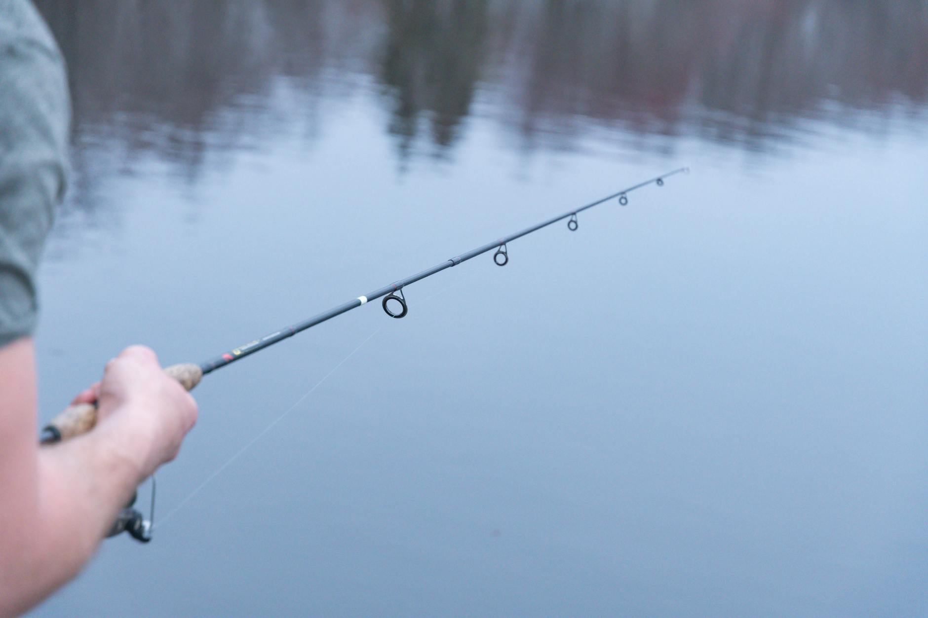 A person holding a fishing rod close-up over calm lake water on a serene day, capturing outdoor leisure.