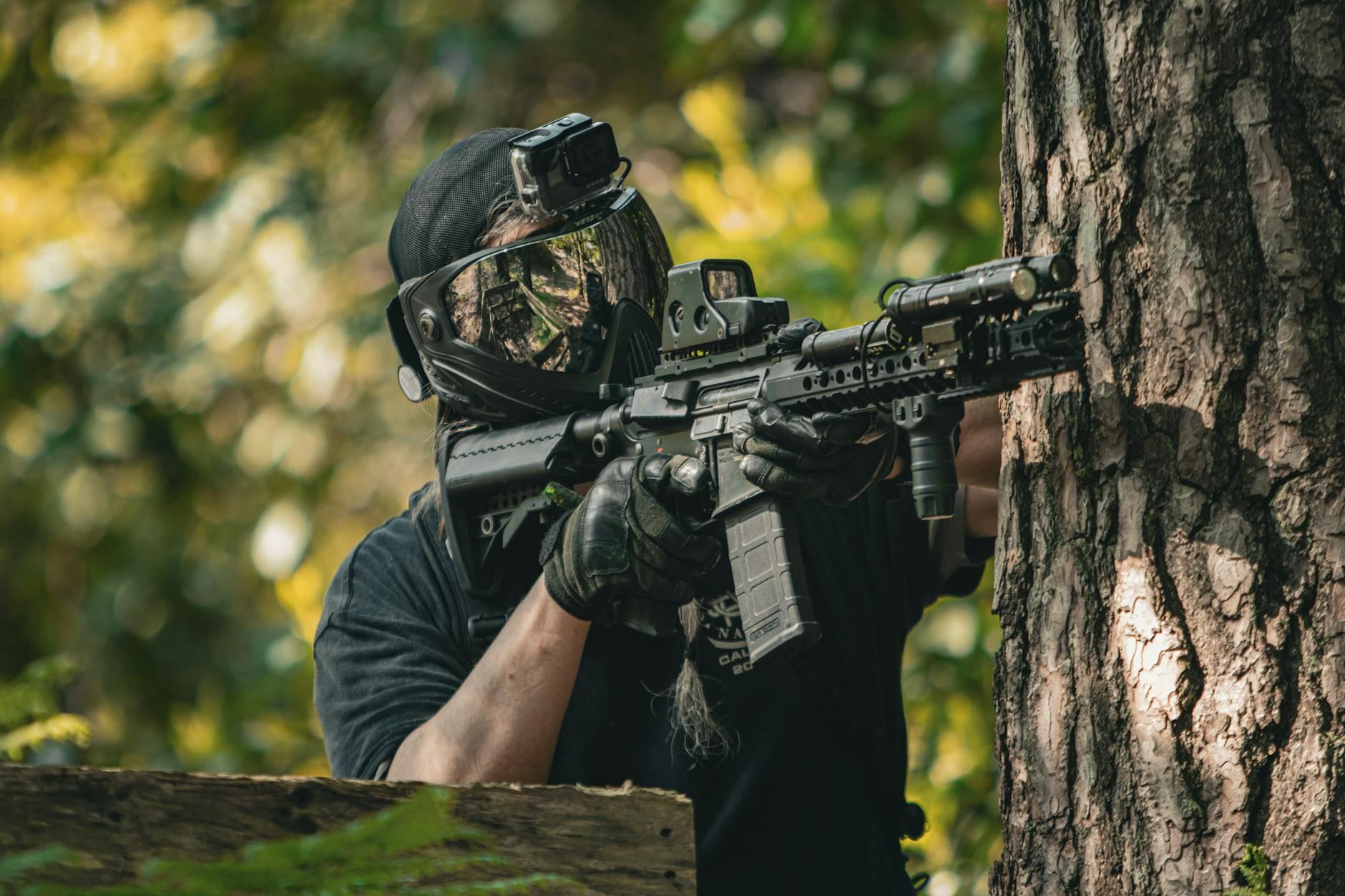 An airsoft player in camouflage crouches behind a tree, focused and ready for action in a tactical game.