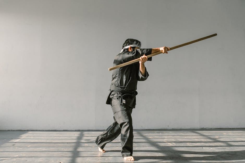 A martial artist practicing Wushu techniques with a wooden staff in a spacious, well-lit studio.