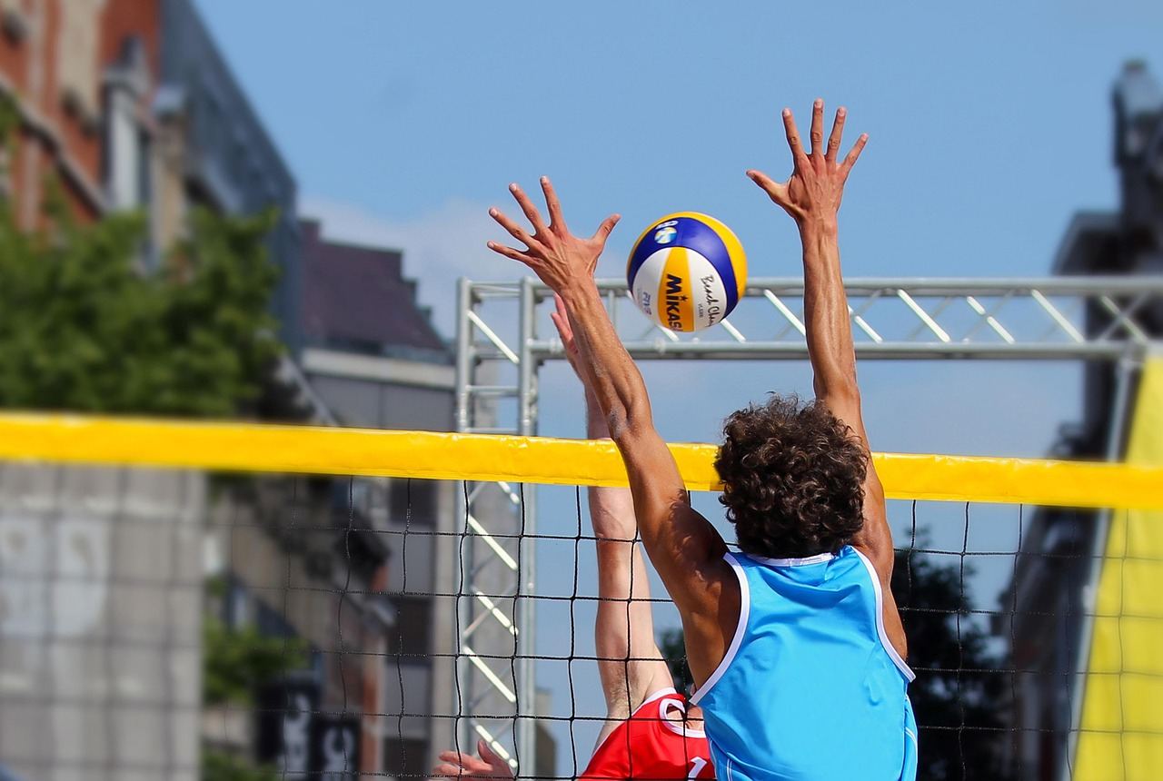 A dynamic scene of players leaping to spike a volleyball over a net on a sunlit sandy beach.