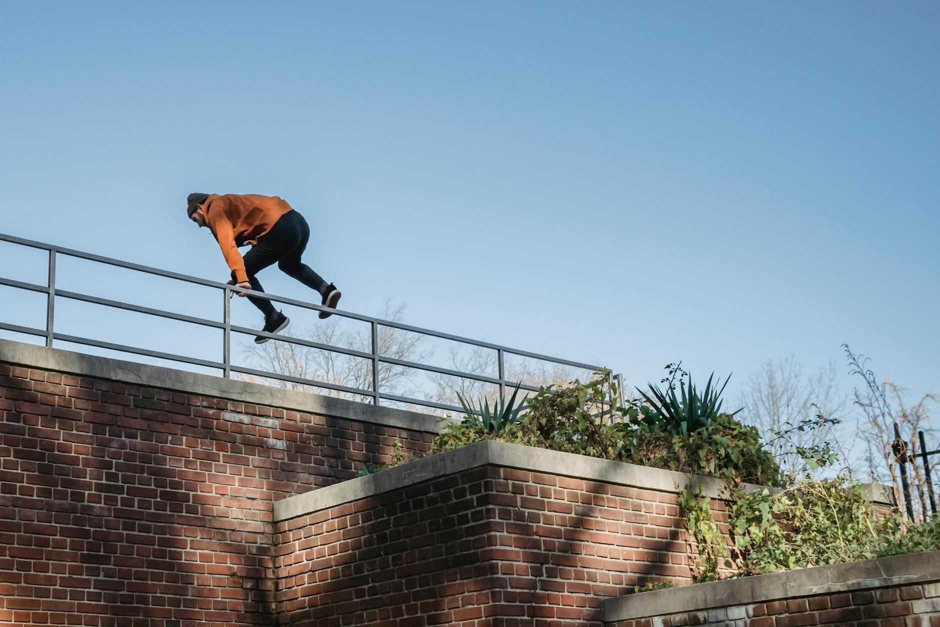 From below side view of anonymous fit male athlete in informal outfit jumping over fence of brick construction in town