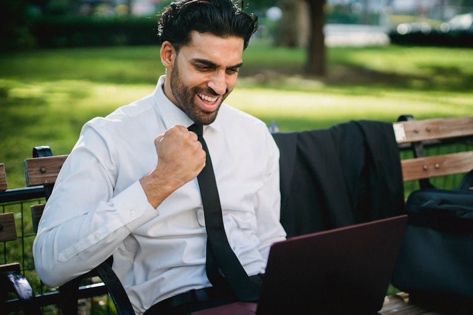 A man in a white shirt and black tie sits on a park bench, smiling broadly as he looks at his laptop. He raises his fist in a gesture of triumph. The background features greenery and a blurred cityscape, suggesting a vibrant outdoor setting conducive to productivity.