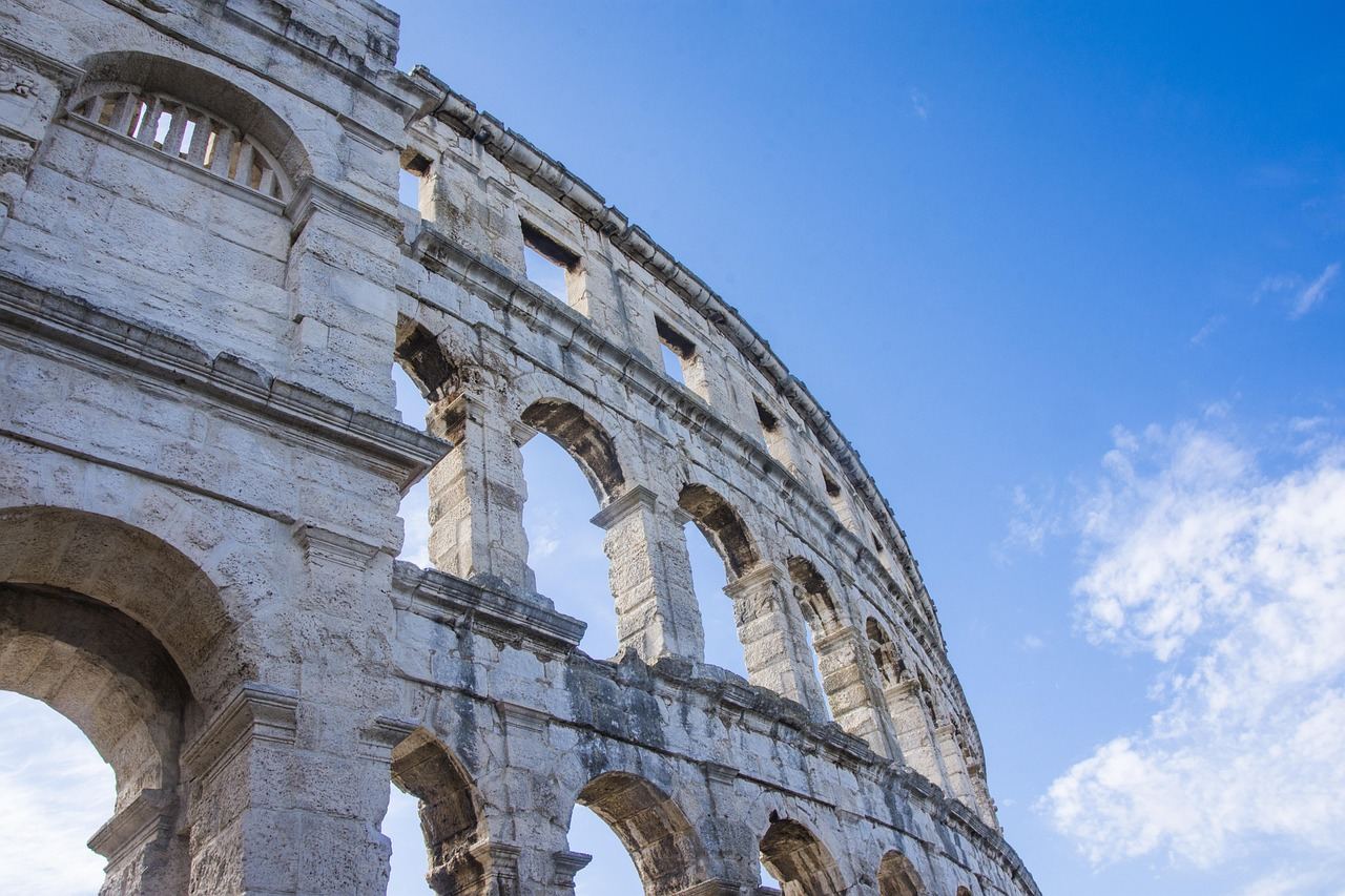 A grand coliseum with tiered seating, surrounded by lush greenery, set against a clear blue sky.