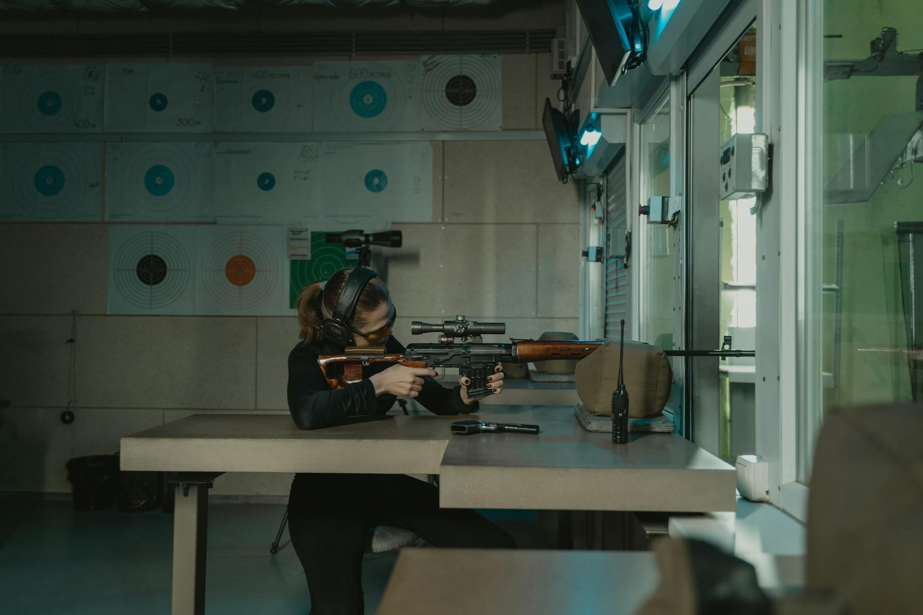 A woman concentrating on target shooting indoors with a rifle, wearing protective gear.