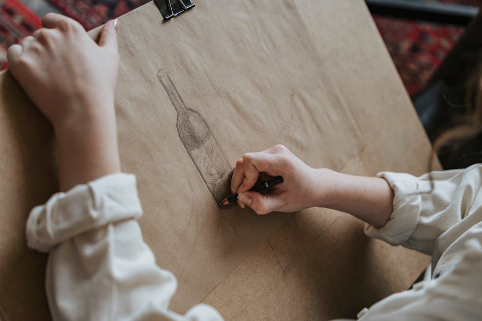 A person with a white shirt is sketching a detailed drawing of a wine bottle on a large sheet of brown paper. The hand grips a pencil, capturing fine details and shading. The background features a patterned rug, adding warmth to the creative space.