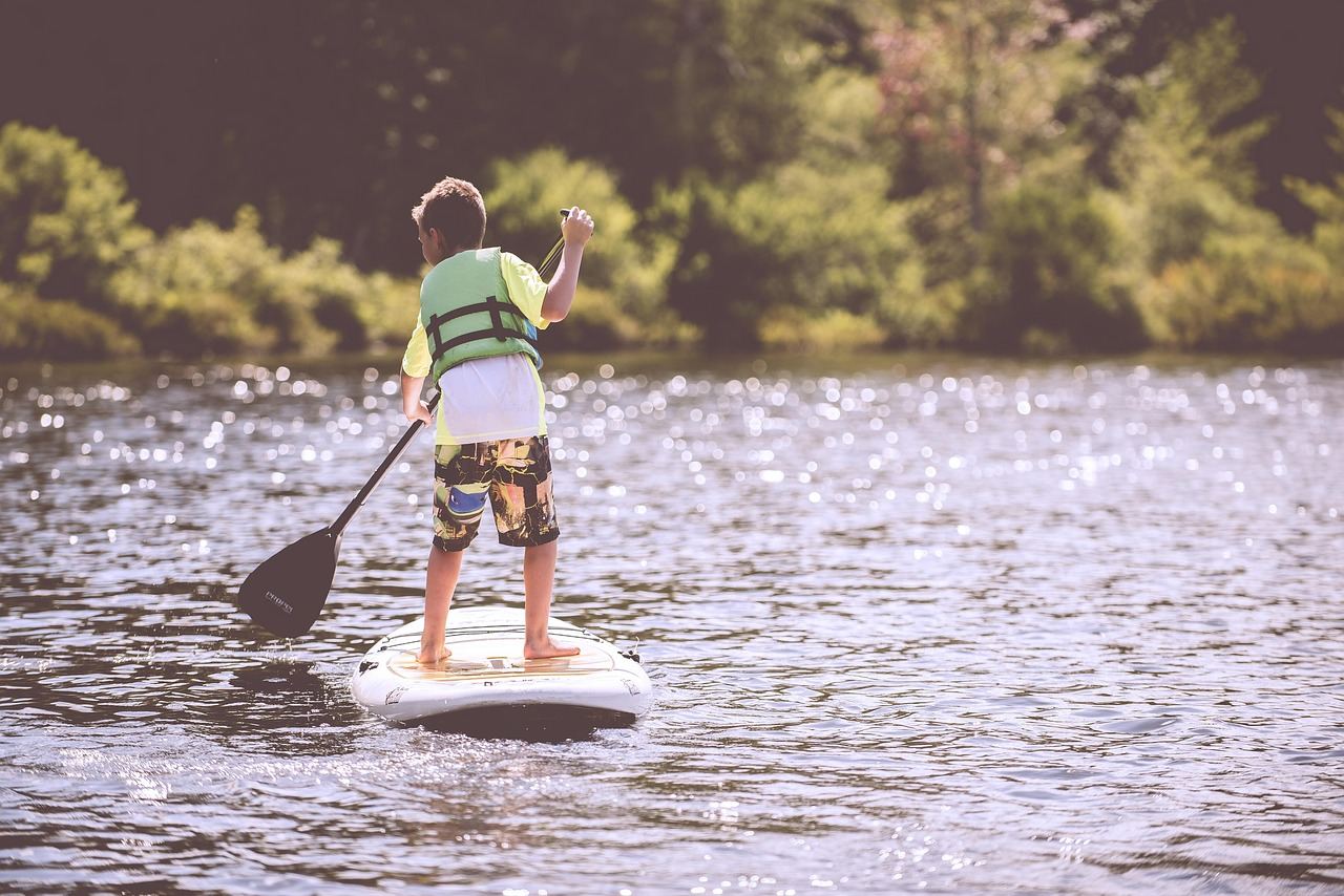 A young boy balances confidently on a bright blue stand-up paddleboard, gliding across calm waters under a clear sky.