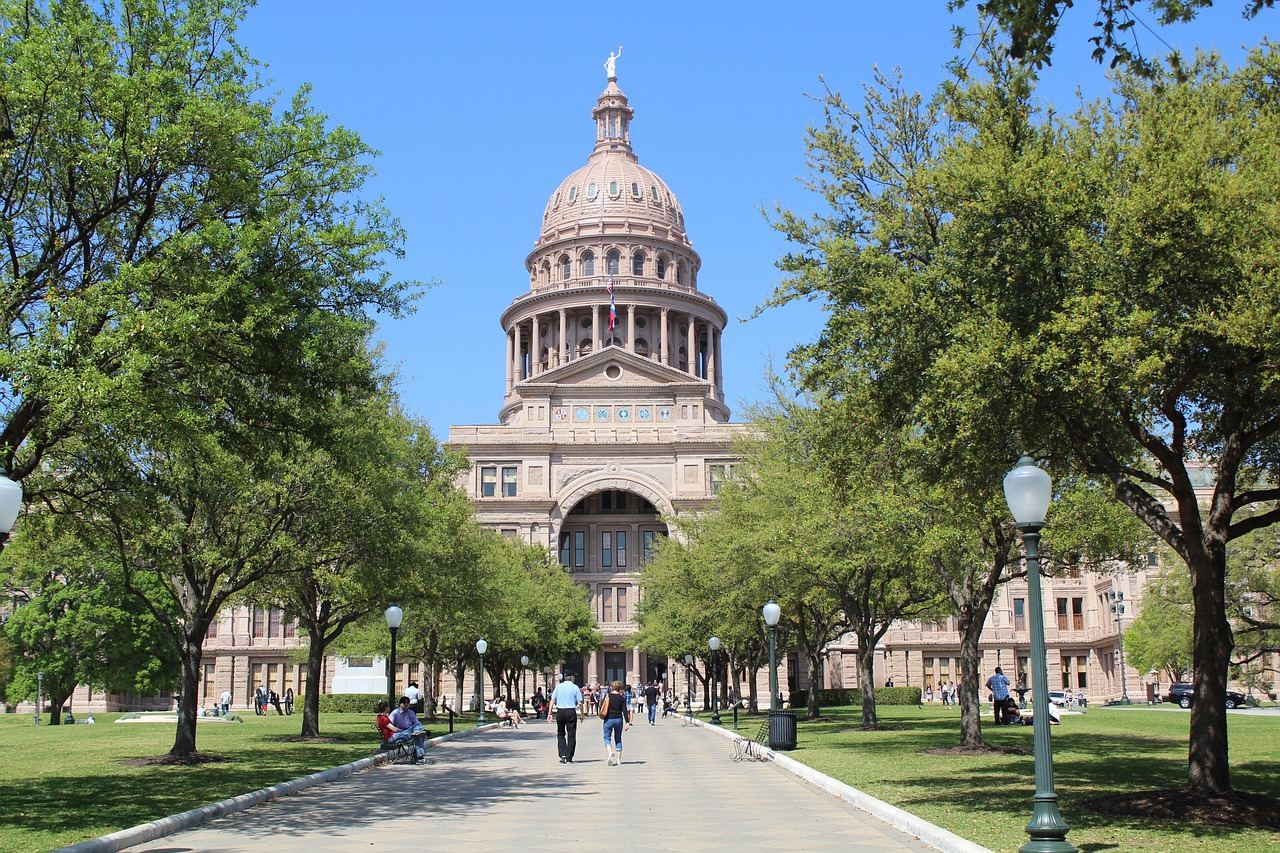 A majestic state capitol building stands proudly against a clear blue sky, surrounded by lush greenery and vibrant flowers.