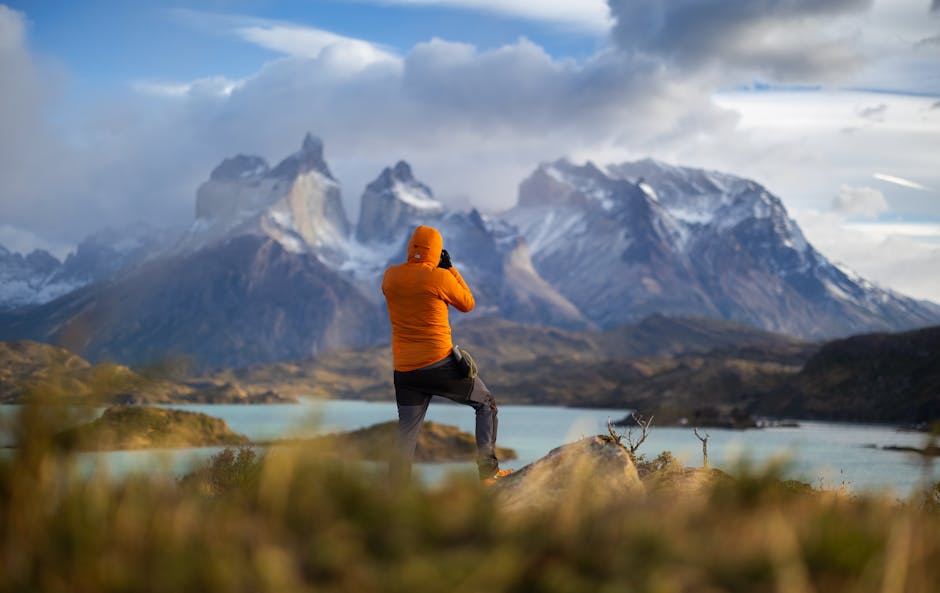 A person in an orange jacket photographs snow-capped mountains and a lake under a cloudy sky.