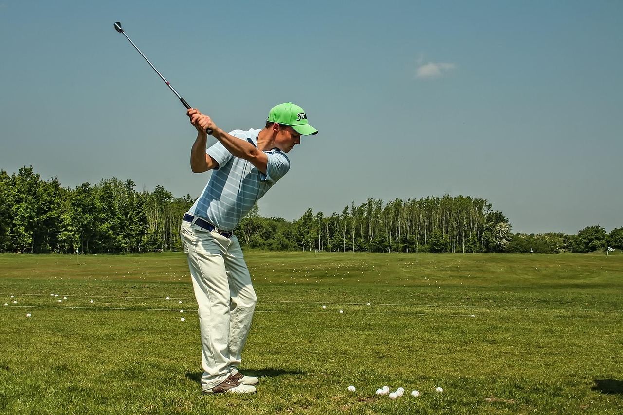 A golfer swings a club, sending a bright white ball soaring against a backdrop of lush green grass and blue sky.