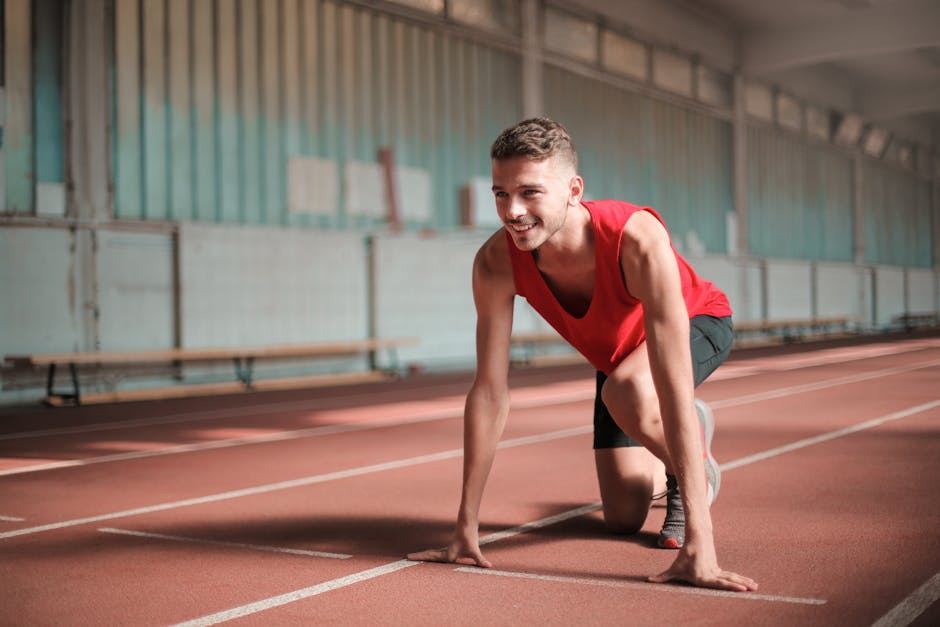 A young man in a red tank top and black shorts is positioned on a running track, ready to start a race. He is crouching with one knee on the ground and a focused, determined expression. The indoor track features marked lanes and a well-lit environment, suggesting an athletic facility.