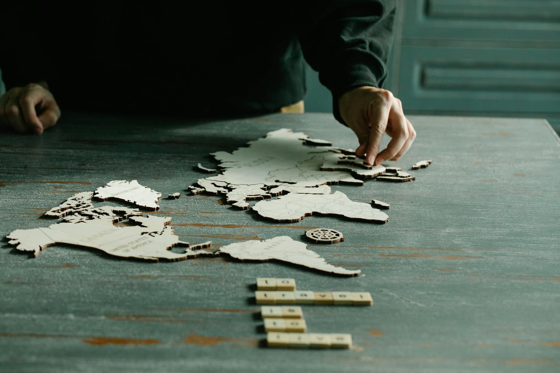 A person carefully fits together pieces of a wooden world map puzzle on a weathered wooden table.