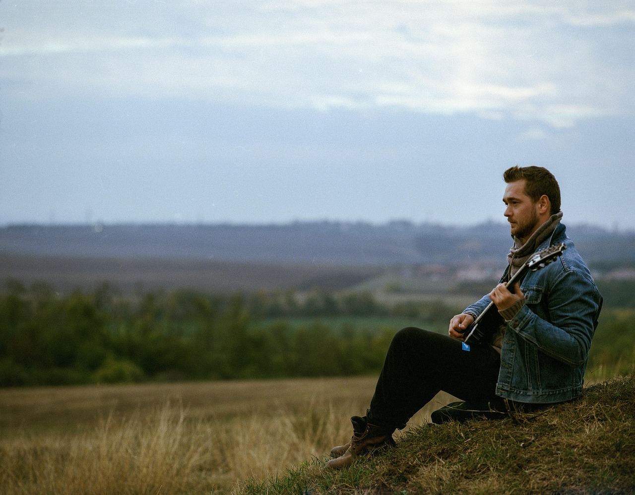 A young man sits on a park bench, strumming a guitar with a focused expression, surrounded by lush greenery.