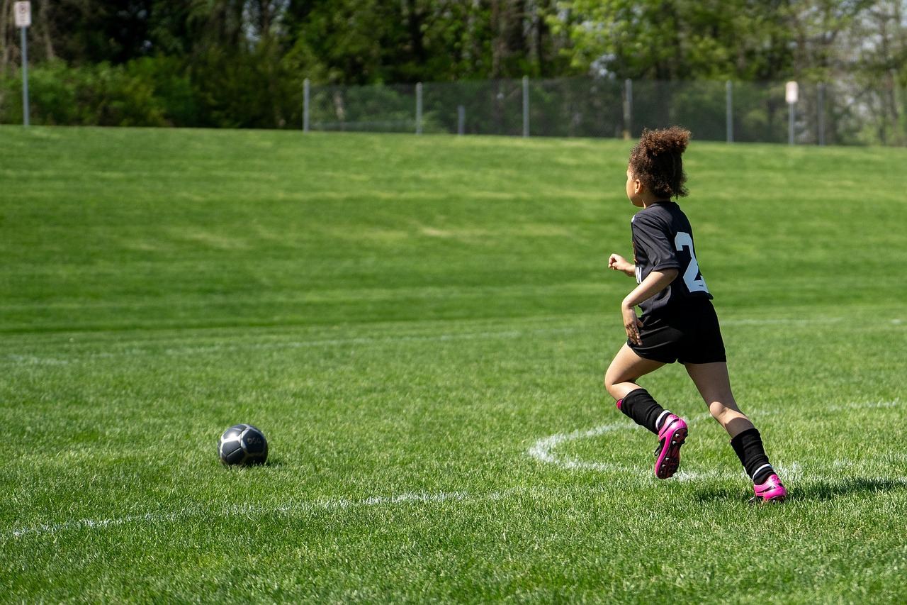 A child wearing a black soccer uniform runs on a green field toward a soccer ball.