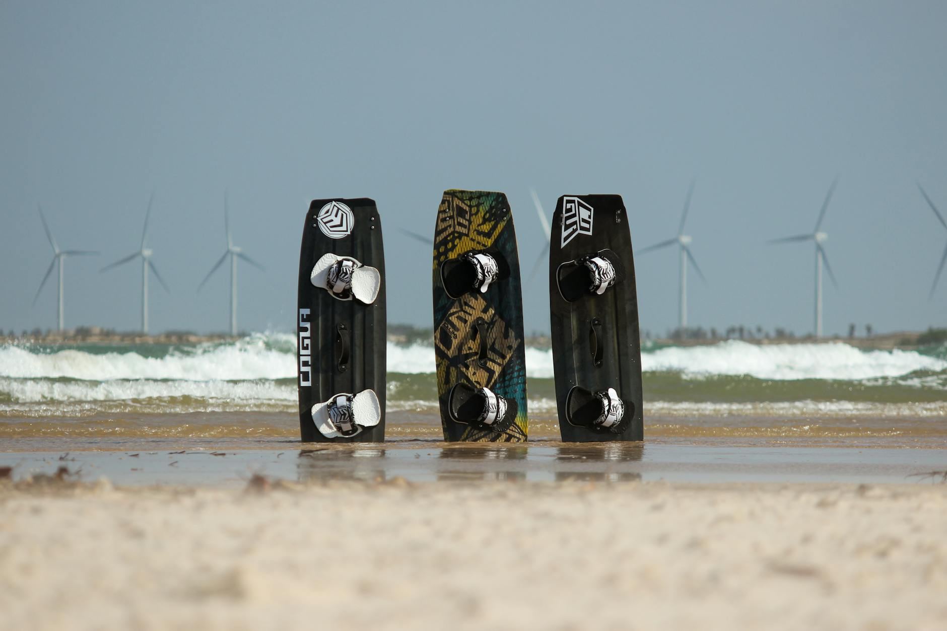 Three vibrant kiteboards rest on a sandy beach, framed by wind turbines against the backdrop of Ceará, Brazil's clear sky.