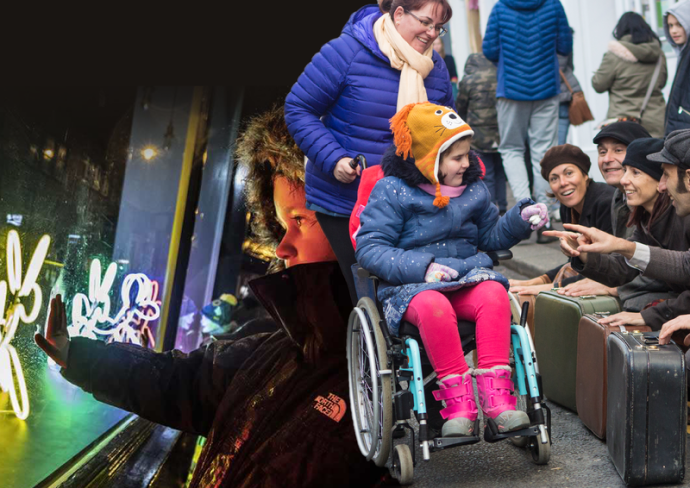 A child touching a window with neon lights dogs on it and another child in a wheelchair talking with people on the street.