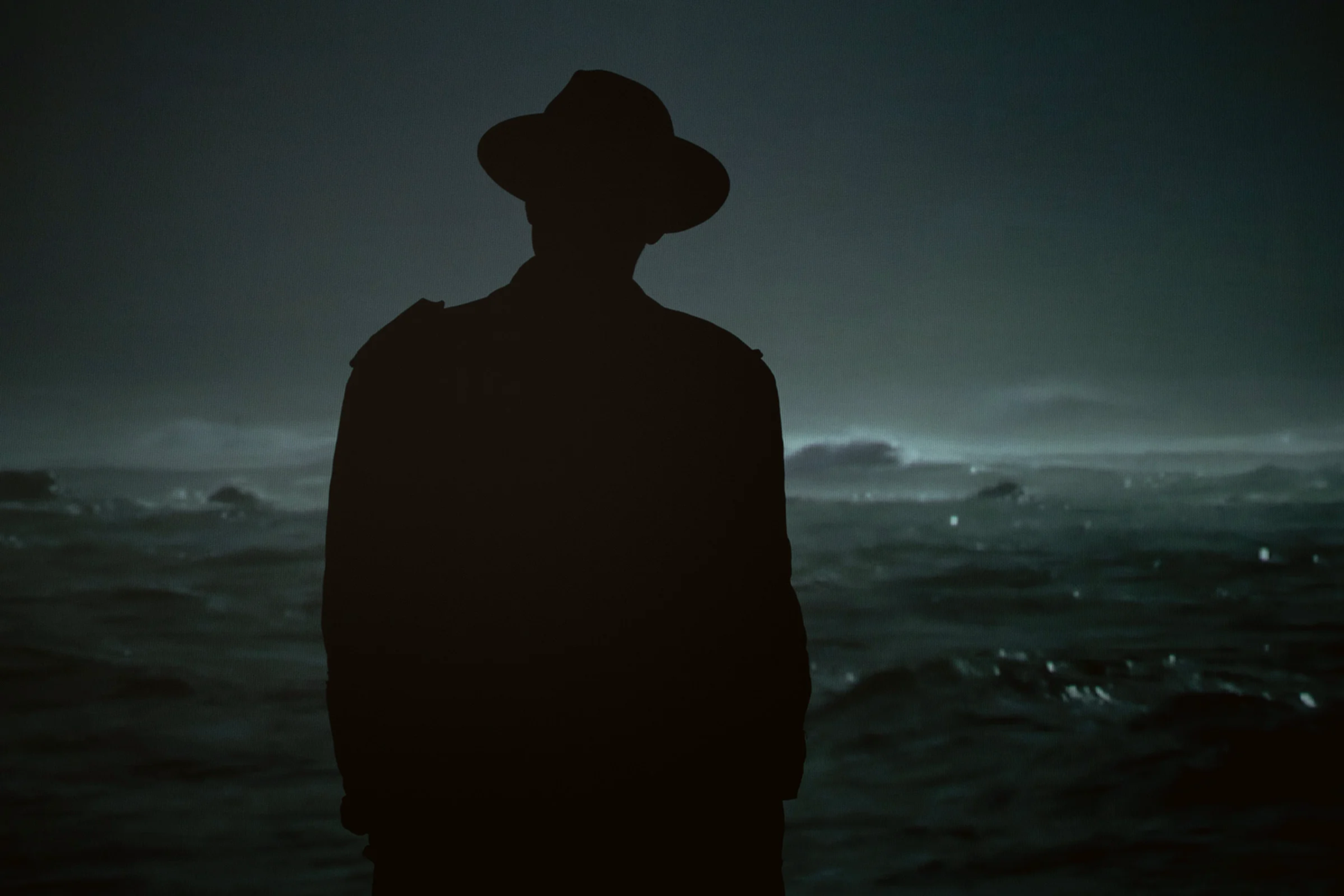 Silhueta de um homem de chapéu, de costas, diante do mar à noite